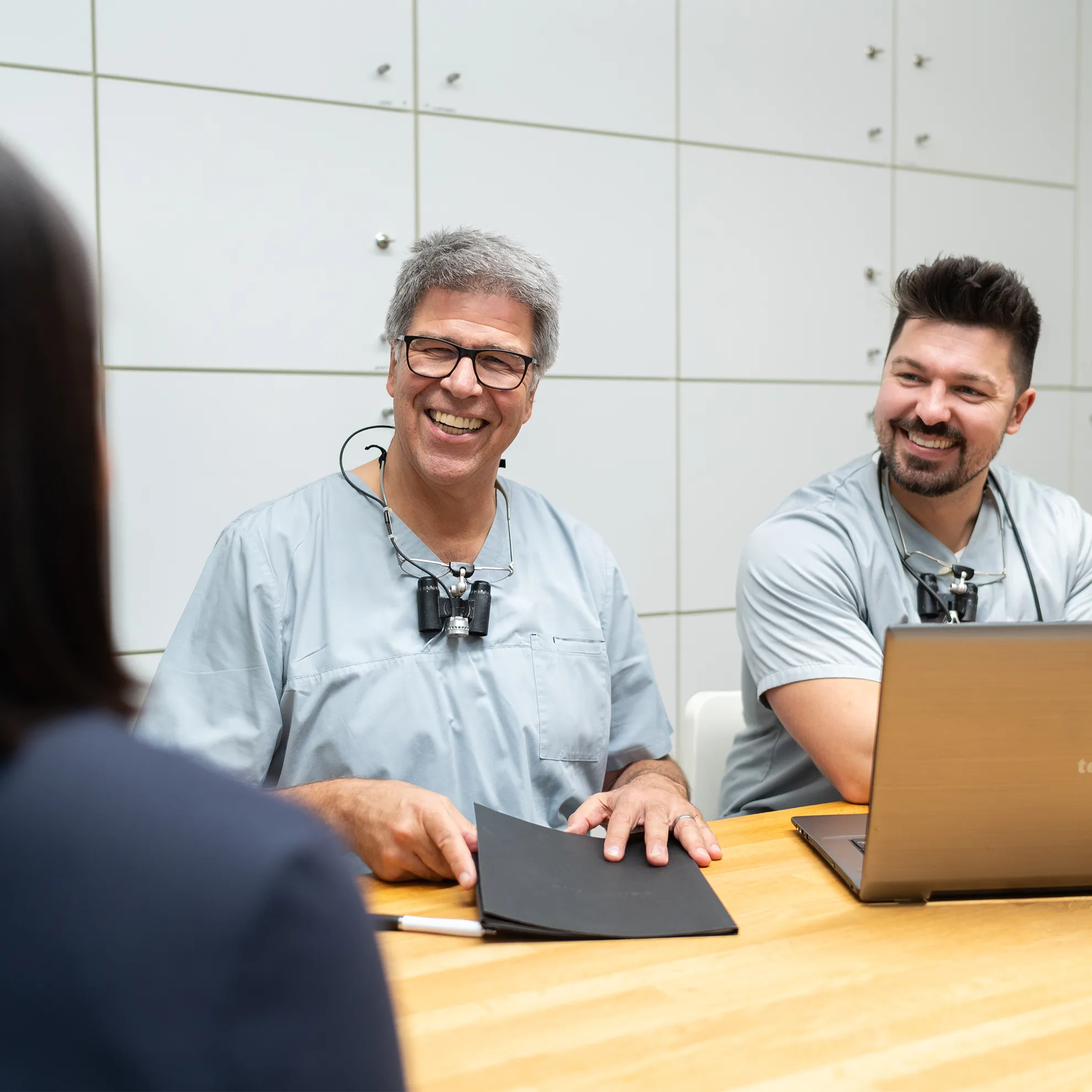 Zwei Zahnärzte sitzen lächelnd am Tisch, während sie mit einer Person sprechen