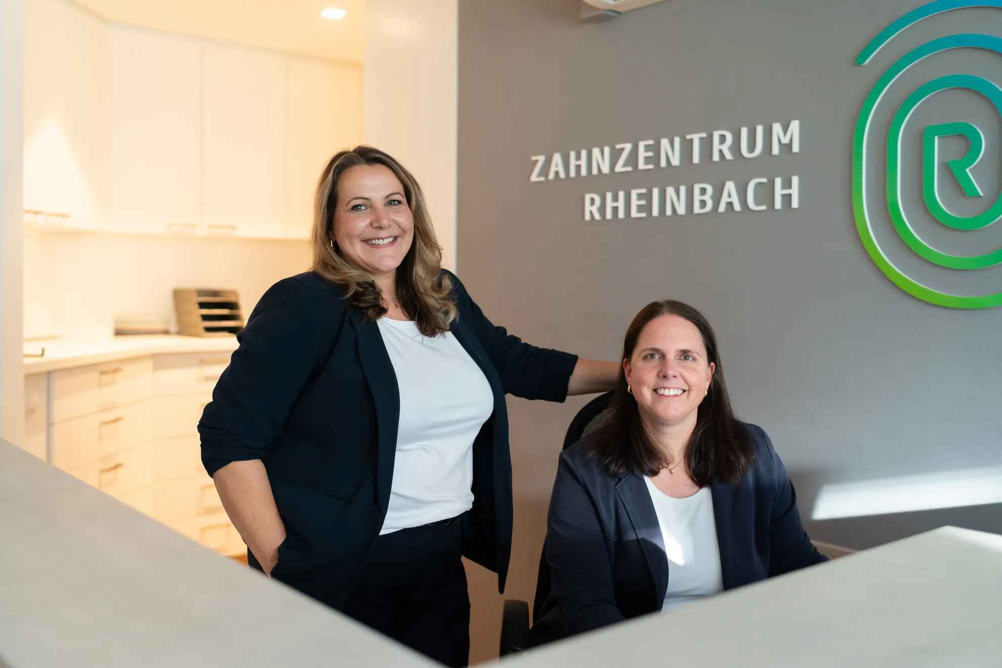 Zwei Damen am Empfang schauen lächelnd in die Kamera, eine steht, eine sitzt